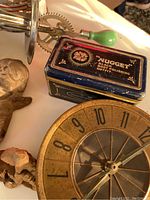 Vintage 'Nugget' black boot polishing outfit tin, decorative clock face dial with one hand and parts of a crank with green handle visible.