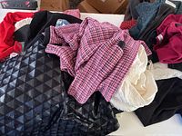 Multiple clothing items displayed on a surface, including a pink and white houndstooth pattern item, black textured velvet skirt, and other garments in plastic bags.
