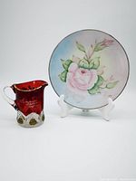 Front view of the ruby stained creamer showing engraved inscription 'Annie 1909' and painted flowers, alongside hand painted floral plate on display stand.