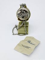 Front view of silver-tone pocket watch with raised motorcycle design on case, hanging on green stand with chain draped.