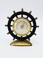 Front view of the ship wheel desk thermometer showing black metal ship wheel with gold-tone rim around thermometer dial.