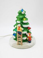 Front view of ceramic Christmas tree with child figurines decorating it. Lights visible. White base simulating snow ground.
