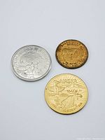 Three coins/tokens placed on a white surface. Left is large silver-colored Tombstone Arizona 1974 medal with engraved cowboy and horse scene. Center bottom is gold-colored 1962 Alaska souvenir money with raised moose and sun. Top right is smaller bronze/copper-colored Green River Whiskey token with text around edge.