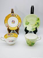 Two vintage teacup and saucer sets displayed upright on stands, left is Royal Paragon with yellow and gold floral designs, right is Aynsley mint green with white floral designs.