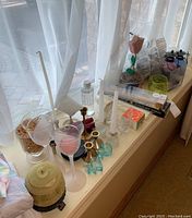 Assortment of candles and candle holders arranged on a windowsill near the sheer curtain. Shows white taper candles, some new in packaging, glass and brass holders, pink frosted glasses, small planters with wood tulip, and a genie oil lamp.