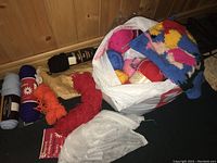 Assorted skeins of yarn in a white and red bag including a colorful knitted Pooh and Piglet project partially visible on top, orange thread, black and white yarn, with some knitting needles.