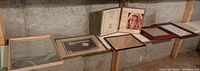 Six photo frames arranged side by side on a wooden railing against a concrete wall, showing a mix of wood finishes and one brass finish frame.