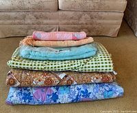 Stack of 6 bedding items showing various colors and patterns including floral, checkered, pastel quilt, and chenille bedspreads with fringe.