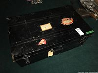 Top and angled view of the rectangular black metal steamer trunk with metal latches and vintage travel stickers showing wear, placed on carpeted floor amid household items.