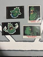 Two rectangular trading cards featuring Slimer on the front with colorful illustrations and captions, shown against a gray background.