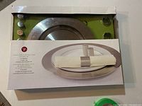 Photo showing the box with the metal charger plate and napkin ring inside, indicating stainless steel material and presentation.