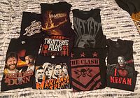 Photo showing 9 black t-shirts with various music-related band graphics and portraits laid out on a bed.