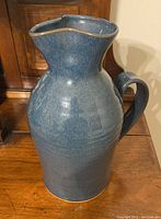 Blue ceramic jug photographed upright on wooden surface against furniture background, showing full shape and handle