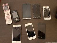 Eight cellphones laid out on table showing front sides, including cracked screens and various models