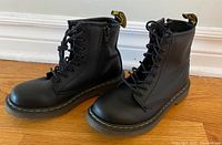 Pair of black leather children's Doc Martens boots photographed from a 3/4 angle, showing lace-up fronts and branded heel tabs.
