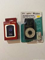 Photo showing the boxed Solutions digital metronome and the Wittner MT60 LED Pendulum Motion metronome in unopened packaging with earphones nearby.