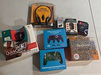 Lot includes Logitech QuickCam Communicate STX webcam in original box, voice music-gaming headset, DexDrive game saver, replacement controller shells, battery pack, and remote control accessories.