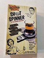 Front side of the Drinkoillo Shot Spinner box showing the shot glass inside the spinner base and usage instructions