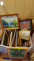 Two bins filled with various framed artworks and needlework. Different wooden frames in light and dark shades. Visible wear and staining.