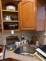 Cabinet open with shelves holding glass pie dish, cake pans, baking dishes, and ceramic ramekins. Countertop has spice rack with glass jars, ceramic mugs, blender, and dish soap by the sink.