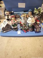 Photo showing assorted Christmas decorations including 12-inch caroler figurines, a snowman, angels, a snow globe, a light-up house, small lamp, and floral holiday arrangements placed on a blue plastic tub.