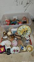 Lot of assorted decorative items including a floral wall clock, multiple vases and bowls with floral patterns, a black and white ceramic cup, and a figurine head, all within a plastic storage bin