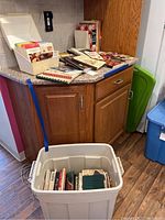 Wide view of cookbooks stacked on countertop and more in a large white plastic bin on floor