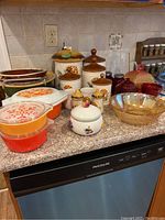 Full lot on countertop showing Pyrex casseroles, mixing bowl, ceramic canisters, white candy dish, clear pitcher, red depression glass sugar and creamer, red depression candy dish, and amber leaf bowl