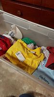Plastic container with various medium-sized shirts in different colors including yellow, green, and blue with visible tags and prints related to Mickey Mouse and Mayberry.
