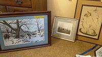 Photo of two framed art prints on floor showing winter scenes, one with stone bridge and skating people
