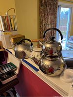 Photo showing a stainless steel ice bucket with handle, a small silver teapot, floral ceramic teapot, and canister sets arranged on a ledge.