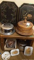 Two octagonal black hand-painted serving trays with floral and fruit designs, one with visible scratches, next to a carved wooden cake plate with lid and a metal cake plate.
