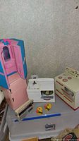 Vintage tin kitchen toys including pink and blue refrigerator, white kitchen sink, and cream-colored oven with red burners shown on a table.