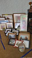 Photo showing various framed wall art of different sizes arranged on floor near wall. Includes different frame styles and art subjects such as floral and portrait.