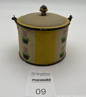 Full front view of the antique metal tin showing hand-painted flowerpot and yellow vertical stripes design, lid fitted on top with small knob, metal carrying handle appears attached on sides