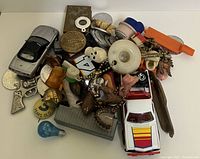 Photo showing a pile of various small items including toy cars, keychains, and charm bracelet pieces, pins, and a small whistle.