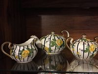 Full view of the Aynsley tea set including teapot, creamer, and sugar bowl with floral patterns and gold accents, displayed on a reflective surface.
