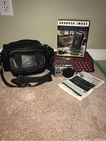 Photo showing the Panasonic Lumix camera, black shoulder bag, boxed Sharper Image electronic key finder, and camera manual on carpeted floor.