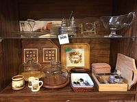 Front view of all items displayed on wooden shelf showing cheese plates with glass covers, terra cotta lidded dish, condiment dish, ceramic pitchers, tile coasters, and other accessories.