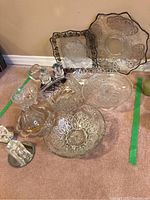 Photo showing assorted clear glass and crystal serving bowls and platters arranged on carpet, various shapes including round, square, scalloped edges with etched floral patterns