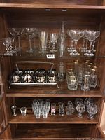 Full view of assorted stemware on top shelf, metal glass caddy holding ombré glasses, multiple tumblers and shot glasses on middle and bottom shelves wooden cabinet