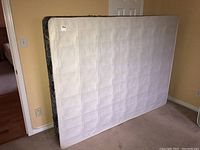 Photo of the box spring standing vertically against a wall showing plain light fabric with stitched square pattern