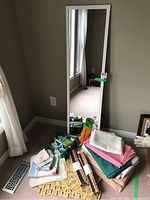 Full view of standing rectangular mirror with white frame beside assorted folded table linens on floor, including pink, teal, and white cloths, quilted table protector, woven placemats.