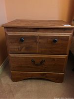 Wooden nightstand with two small top drawers and one large lower drawer, black knobs on small drawers, metal handle on large drawer, natural wood grain finish
