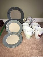 Photo showing ceramic dinner and salad plates with floral design, mugs with leaf designs, and tea pot with matching cup on carpet.