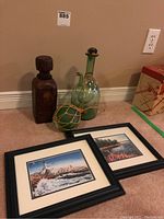 Two framed and matted nautical prints lying on the floor with three glass decorative items: green glass decanter, green glass ball wrapped in rope net, and a dark glass liquor bottle wrapped in leather-like material.