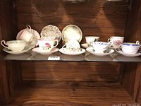 Shelf display of 11 assorted vintage teacups and saucers with floral and gold trim patterns.