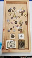 Top view of wooden tray displaying a variety of foreign coins spread out, showing different sizes and metals. Two paper currency notes and two large coins in cardboard sleeves are placed at the bottom.