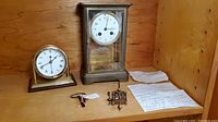 Two desk clocks side by side on a wooden surface including brass mantle clock and nautical desk clock with papers and keys beside them.