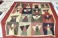 Full view of handmade quilt blanket showing fifteen angel blocks arranged in three rows of five, each angel with distinct fabric patterns in muted earth tones, white halos, and simple geometric designs.